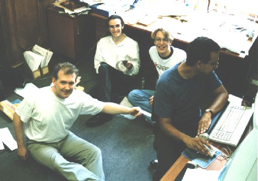 A photo from 1996 showing four people in an office setting with papers scattered around: Jim Burns and Adam Donlin, both PhD students at the time, myself looking very young, and Satnam Singh at a desk trying to focus on work.