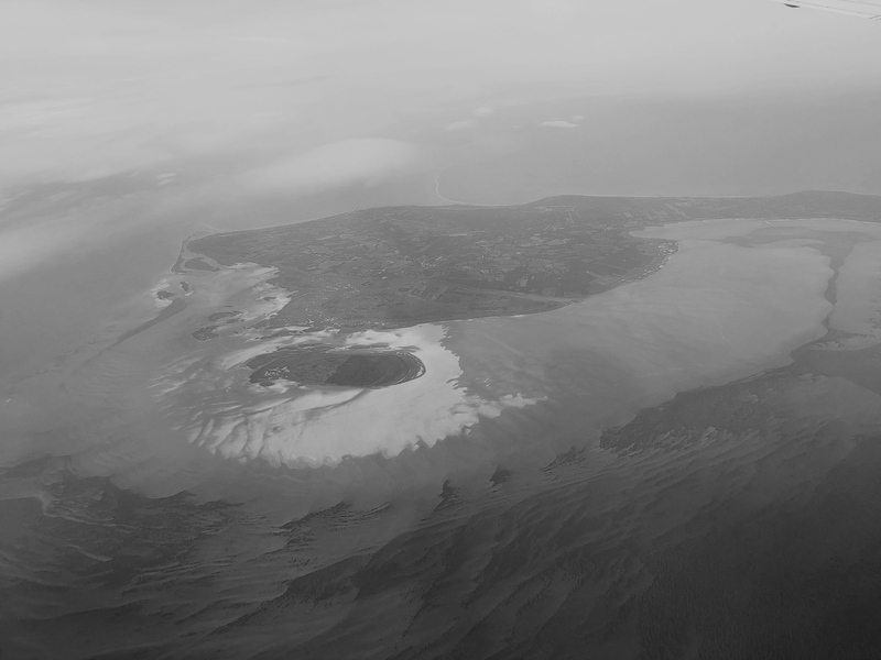 A photo of an island in a sea taken from the window of an airplane. The island has a swirly shape, which is emphasised by the sand banks around it.