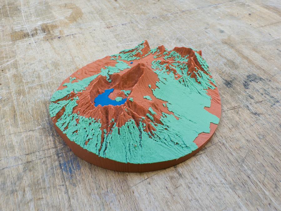 A photo of a 3D printed circular topographical model sitting on a wooden surface. The model depicts a mountainous volcanic landscape with a crater lake, using a colour-coded system where red/orange represents the ground, green shows forested areas on the side of the volcano, and blue depicts a crater lake at the centre.