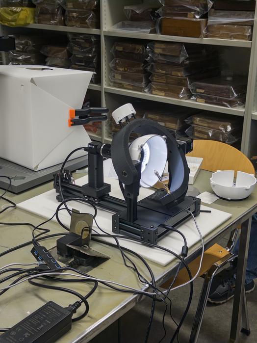 Photo of an open source scAnt 3D scanner setup on a laboratory workbench, featuring a black motorised scanning apparatus with a white circular platform, surrounded by various cables and electronic components.