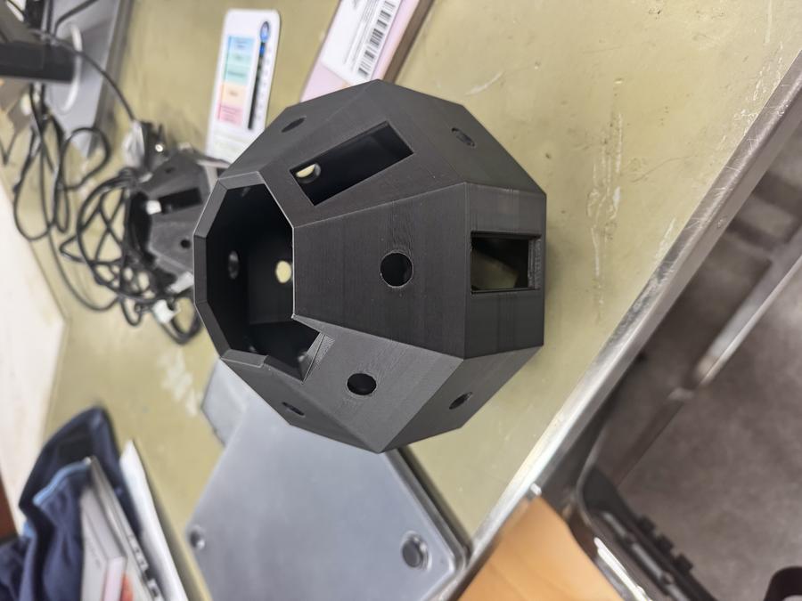 Photo of a black 3D-printed dodecagonal (12-sided) jig with multiple circular holes and rectangular openings, designed as a prototype for positioning insects during mobile phone scanning, sitting on a laboratory workbench with various equipment and papers visible in the background.