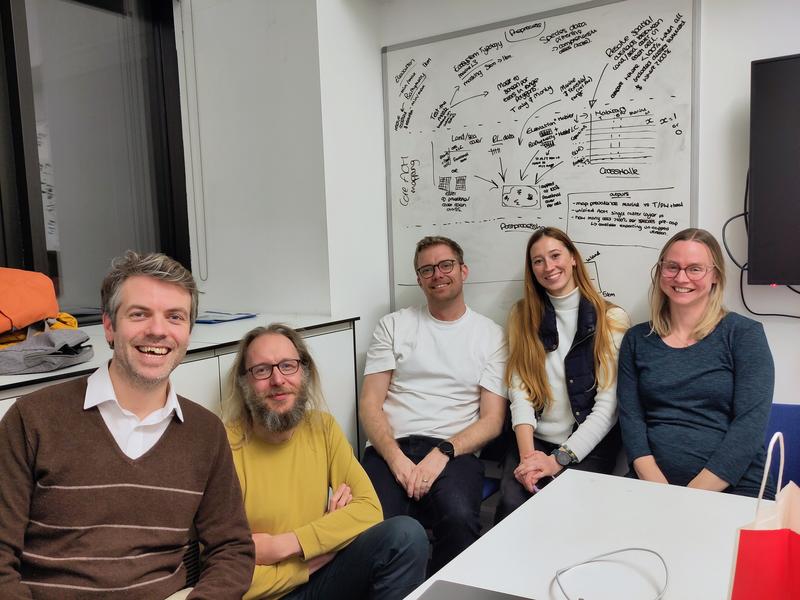 A photo of five people sitting together in an small meeting room, smiling at the camera, with a large whiteboard covered in diagrams and notes visible on the wall behind them and a desk with papers in the foreground.