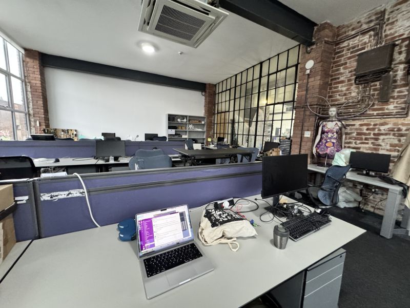 A photo of a laptop on a desk in a large brick room with many unoccupied disks in it. To once side is a mannequin wearing a dress made from re-used sweetie boxes and with angel wings made from old bike wheels.