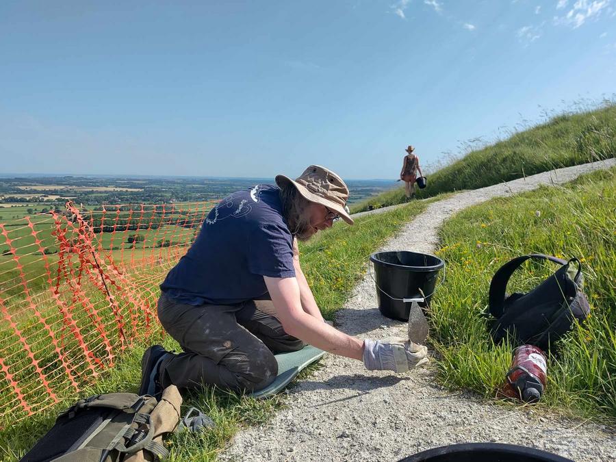 A photo of me kneeling next to a row of chalk in a grassy hillside. I'm using the end of the handle on a trowel to tamper down the chalk after removing a weed.