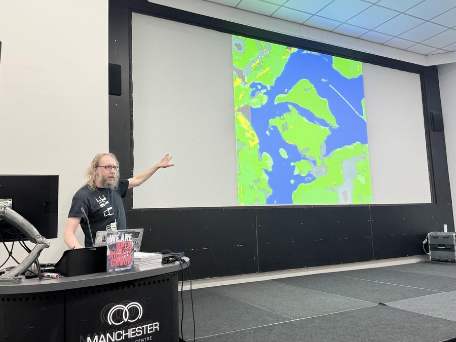 A photo of a person with long hair gestures towards a large screen displaying a colourful map visualization.